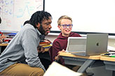 A teacher helping a student on a laptop. Links to Gifts of Life Insurance